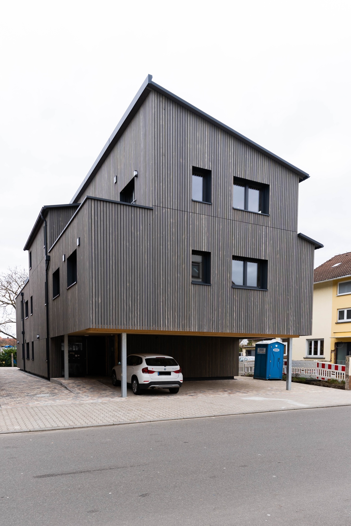 Ein modernes zweistöckiges Gebäude mit Holzverkleidung, einem offenen Garagenbereich im Erdgeschoss und einem weißen Auto darin.